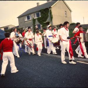 Children's Oss procession walking along pavement with boy teasing Blue Ribbon oss