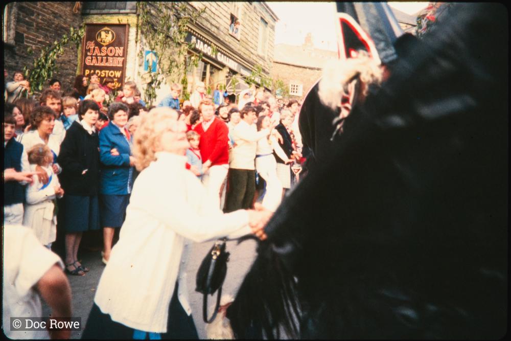 Woman holding hands with Old Oss under maypole, Blue Ribbon oss dancing in background