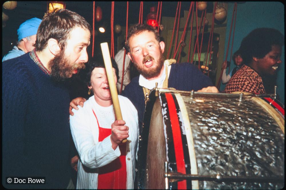 Woman playing bass drum, with 2 men singing