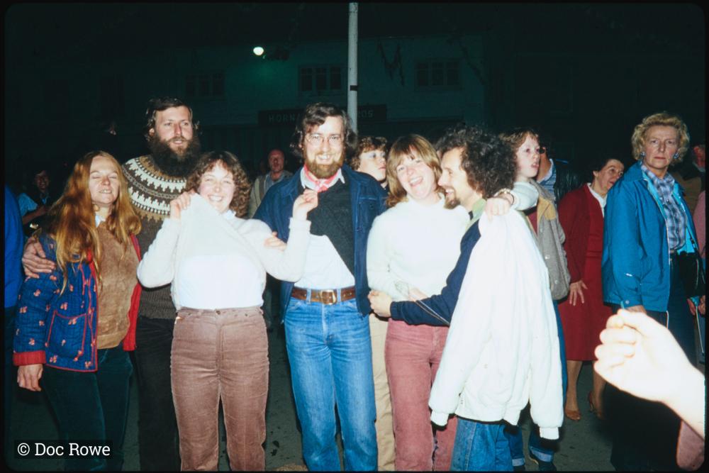 Night time, people smiling and pulling up jumpers under Maypole